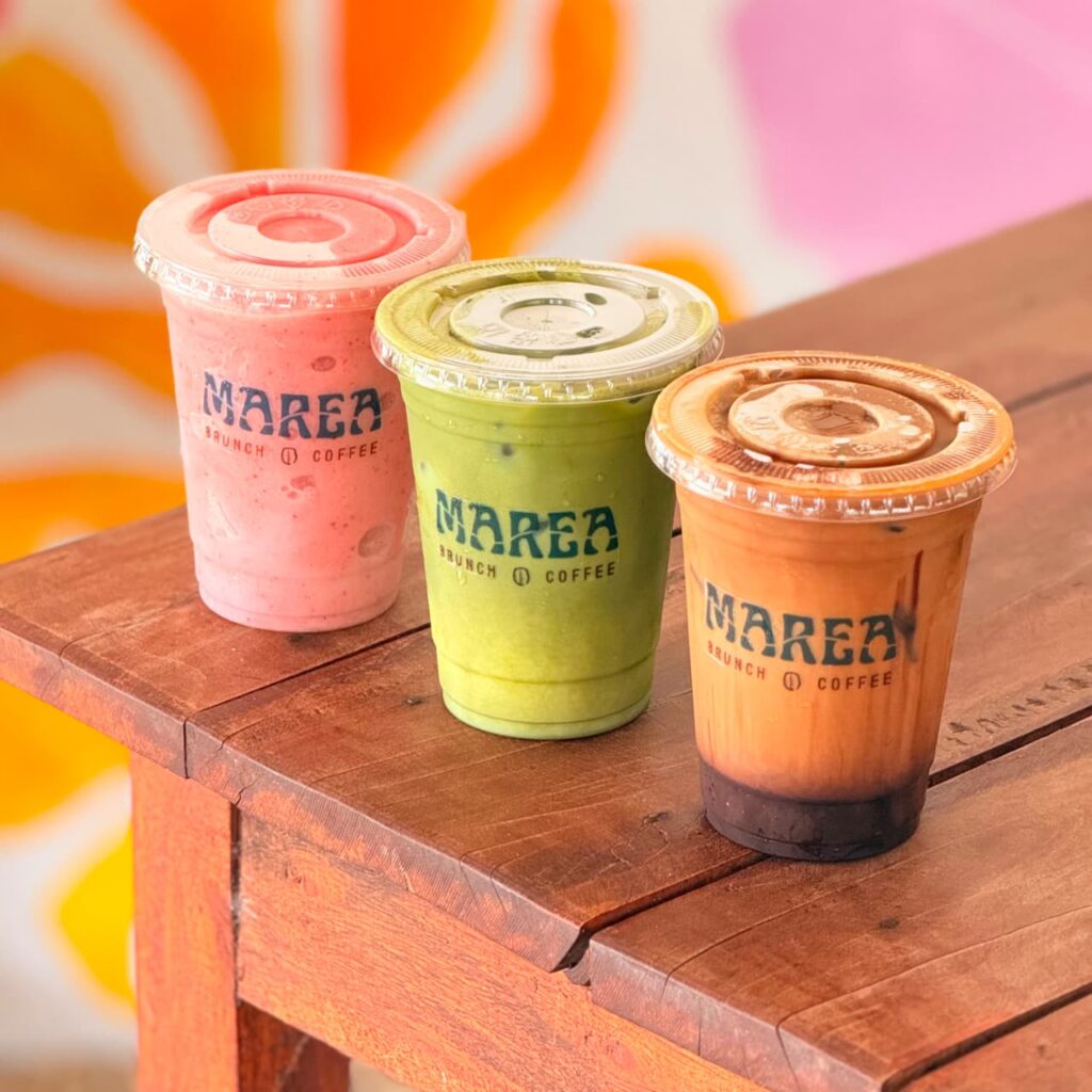 Tres bebidas frías de Marea en vasos con el logotipo, sobre una mesa de madera: una rosa, un matcha y una de café con capas