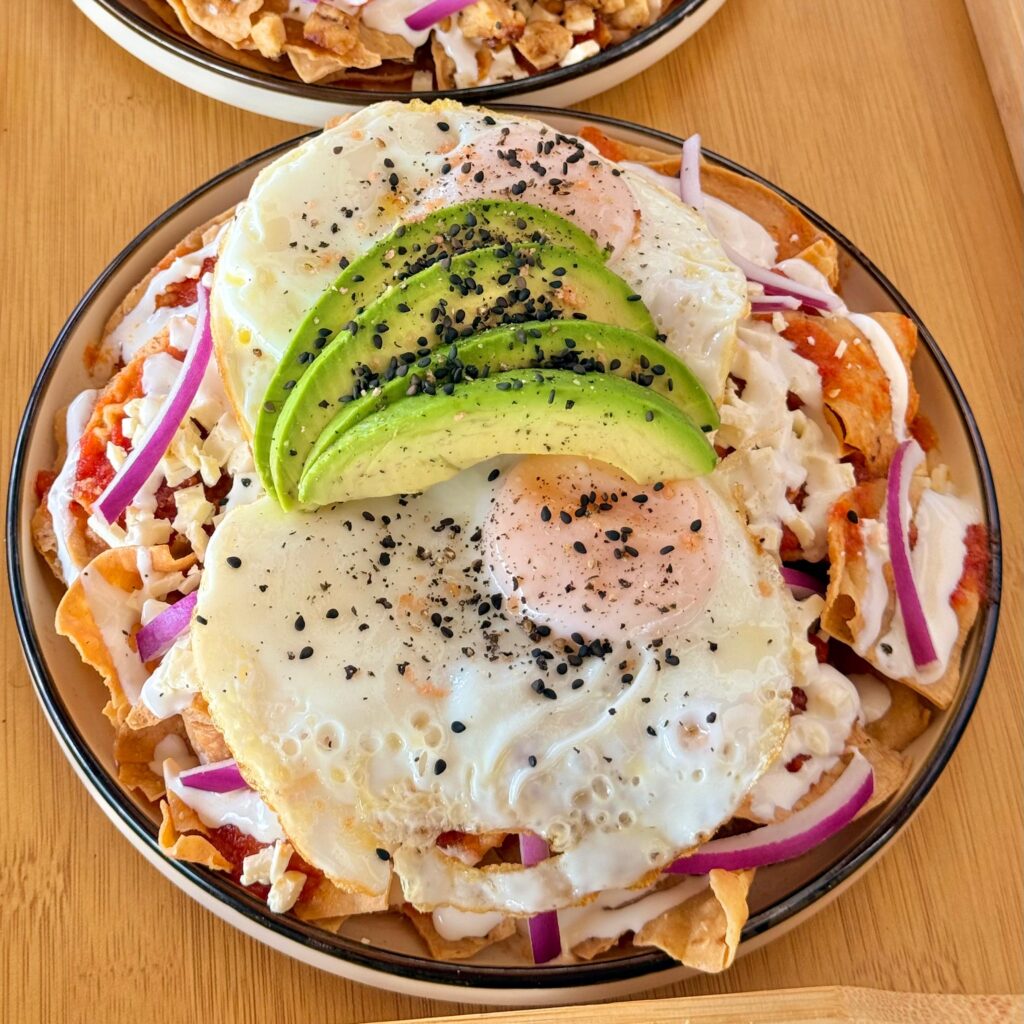 Plato de chilaquiles rojos con huevo estrellado, aguacate en rebanadas, crema, queso y cebolla morada